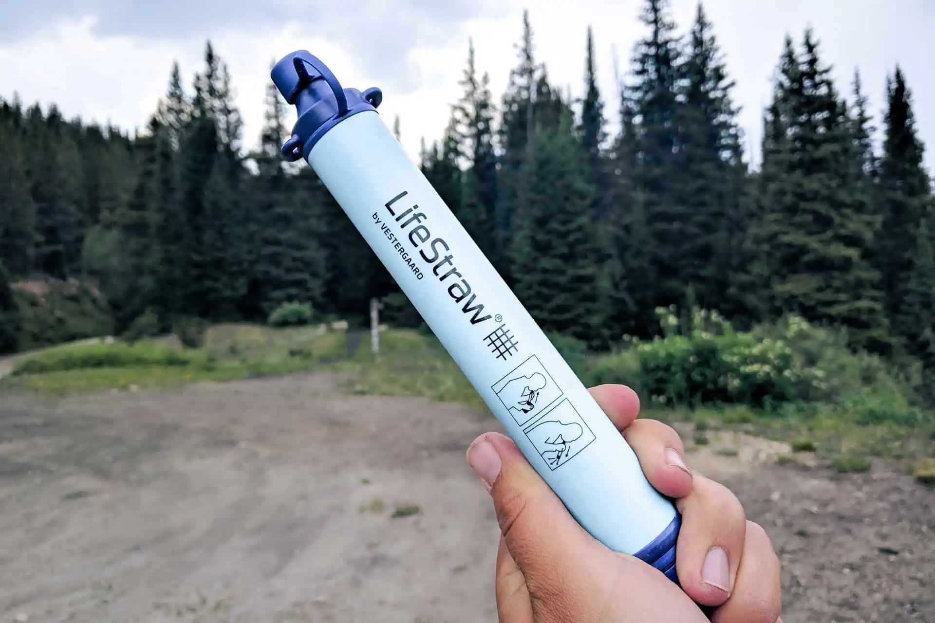 A person holds a LifeStraw with a forest backdrop, showcasing the portable blue water filter ideal for outdoor adventures. Its label features pictorial instructions, ensuring easy use in natures embrace.