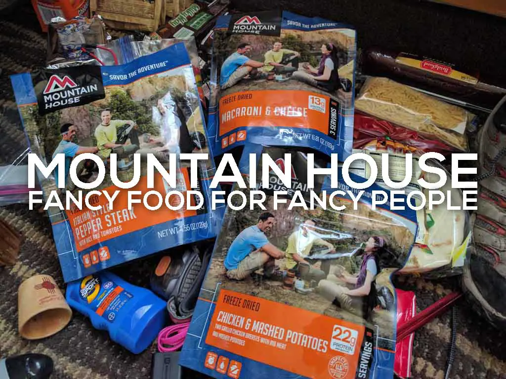 Packets of Mountain House freeze-dried food, including Pepper Steak, Macaroni & Cheese, and Chicken & Mashed Potatoes, are displayed. The text on the image reads Mountain House Fancy Food for Fancy People. Various camping supplies are visible around the packets.