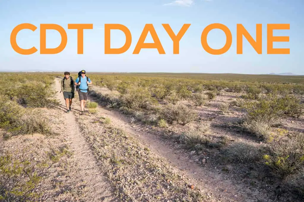 Two hikers traverse a desert trail beneath a clear blue sky. The flat terrain is adorned with sparse vegetation. Large orange text declaring CDT DAY 1 dominates the top of the image.