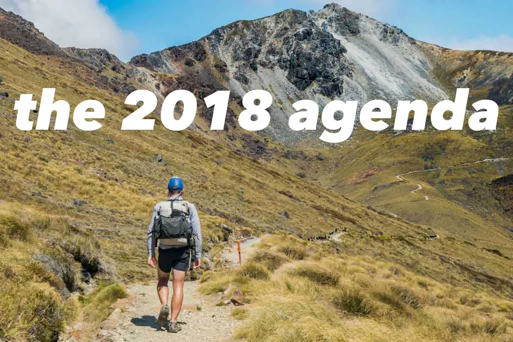 A hiker wearing a backpack and cap walks up a dirt path through grassy hills towards a rocky mountain. The sky is partly cloudy. Overlaid text reads Whats On The 2018 Agenda.