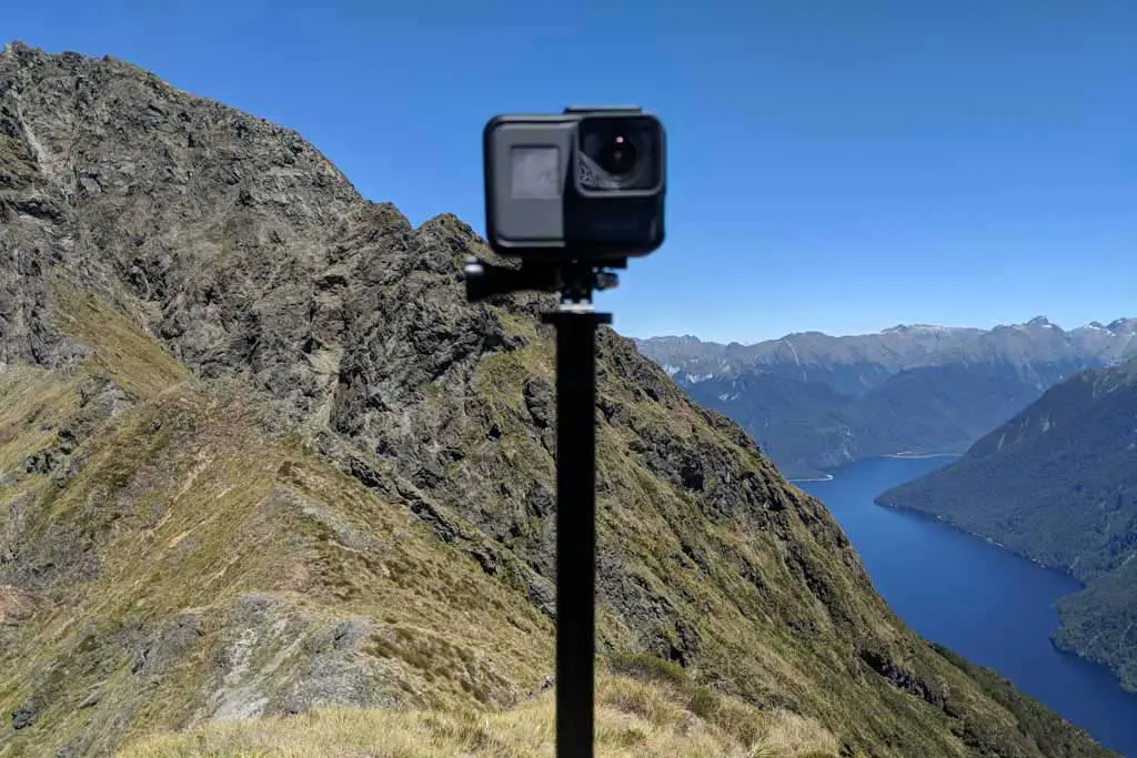 A GoPro camera mounted on a pole, with a scenic background of rugged mountains and a blue lake under a clear sky.
