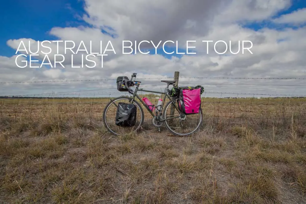 A bicycle loaded with touring gear stands by a barbed wire fence on a grassy plain under a cloudy sky. Text on the image reads Australia Bike Touring Essentials.