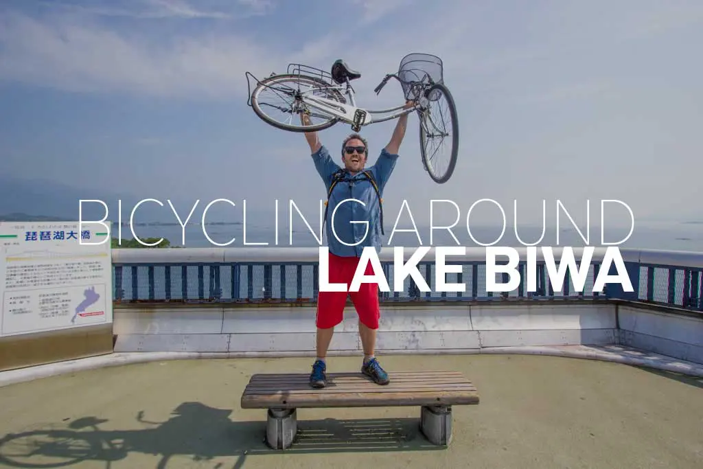 A person stands triumphantly on a bench, raising a bicycle overhead at the scenic overlook of Lake Biwa, water glistening in the background. The text reads Bicycling Around Lake Biwa.