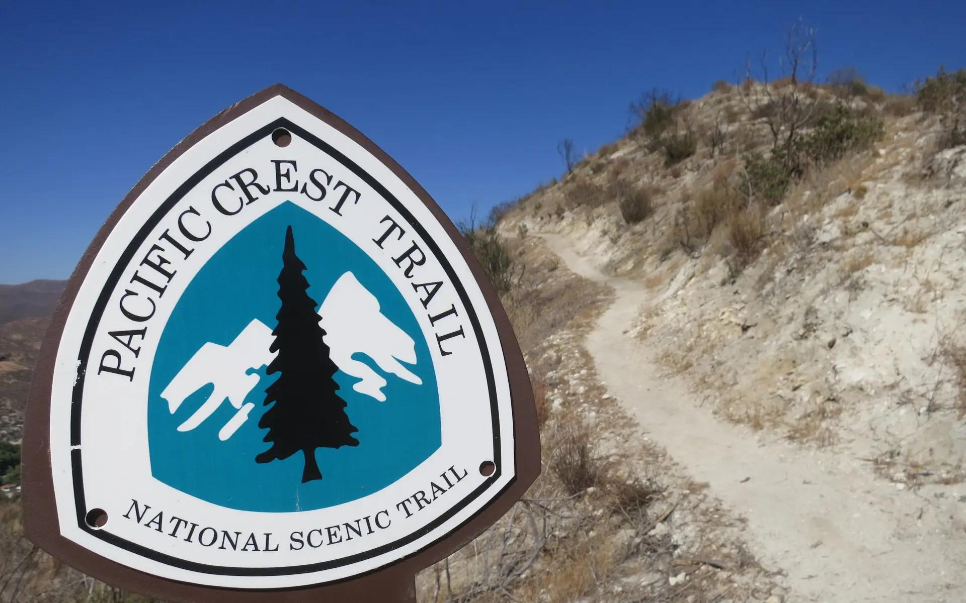 A sign for the iconic Pacific Crest Trail, adorned with mountain and tree graphics, stands near a dirt path winding through the dry, hilly landscape under a clear blue sky.