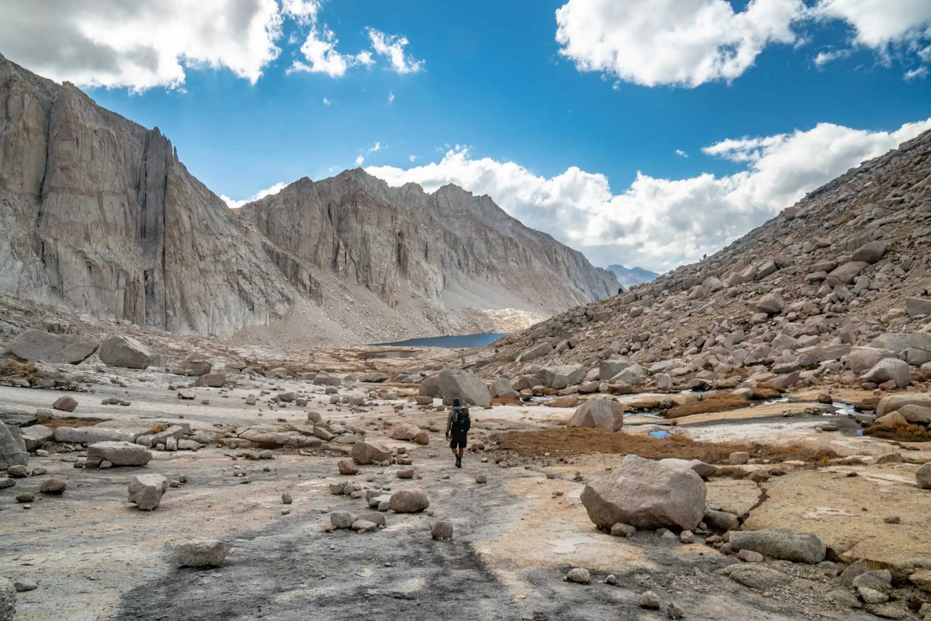 A lone hiker, equipped with pct gear, walks along a rocky path surrounded by rugged mountains under a partly cloudy sky. A distant lake is visible, bordered by steep cliffs. The scene conveys a sense of solitude and natural beauty.