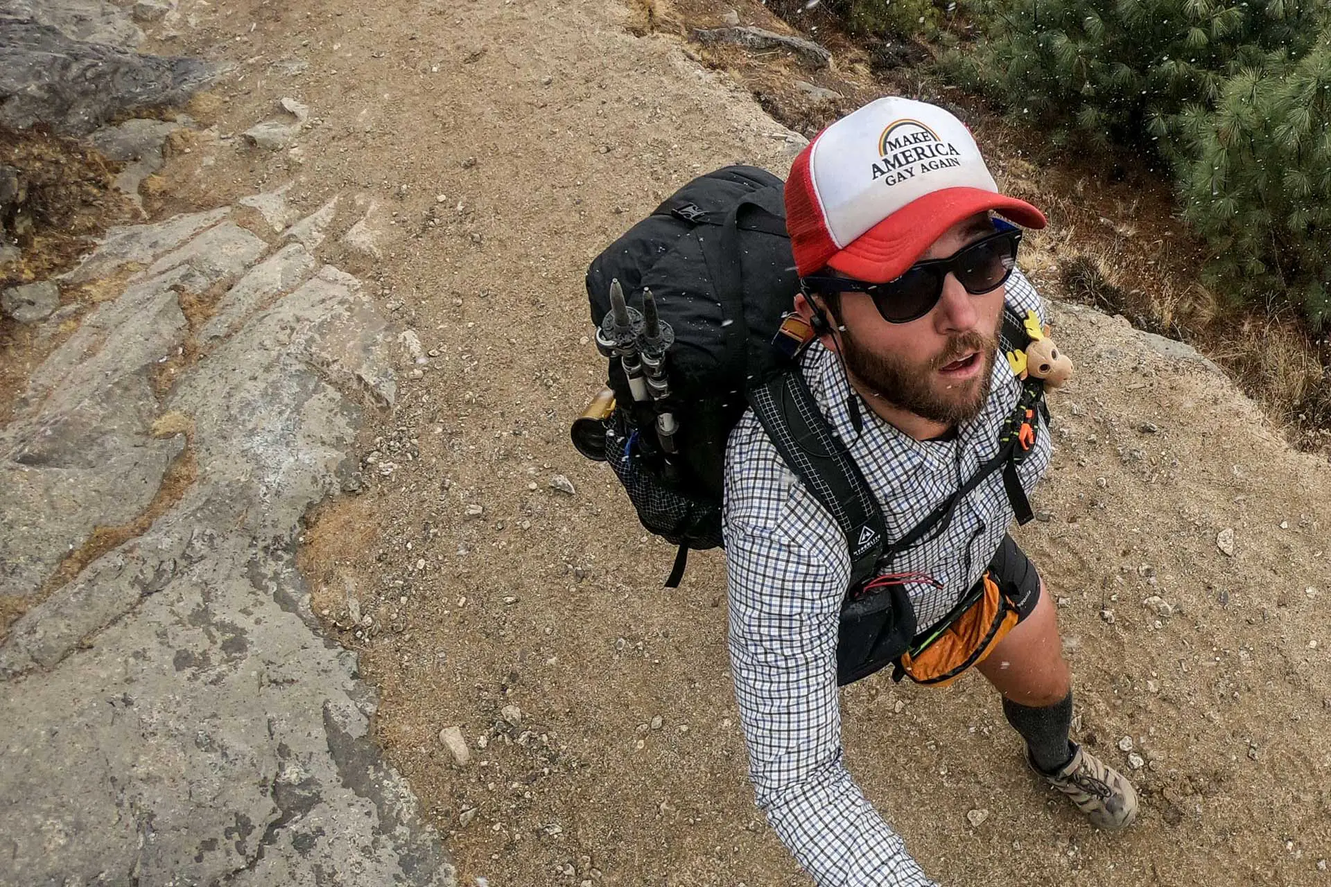 A man with an adventurous agenda strides confidently on a rocky trail, sporting sunglasses, a red and white cap, and hiking gear. His large backpack and poles help navigate the dry path dotted with rocks and sparse vegetation, as seen from an aerial view.