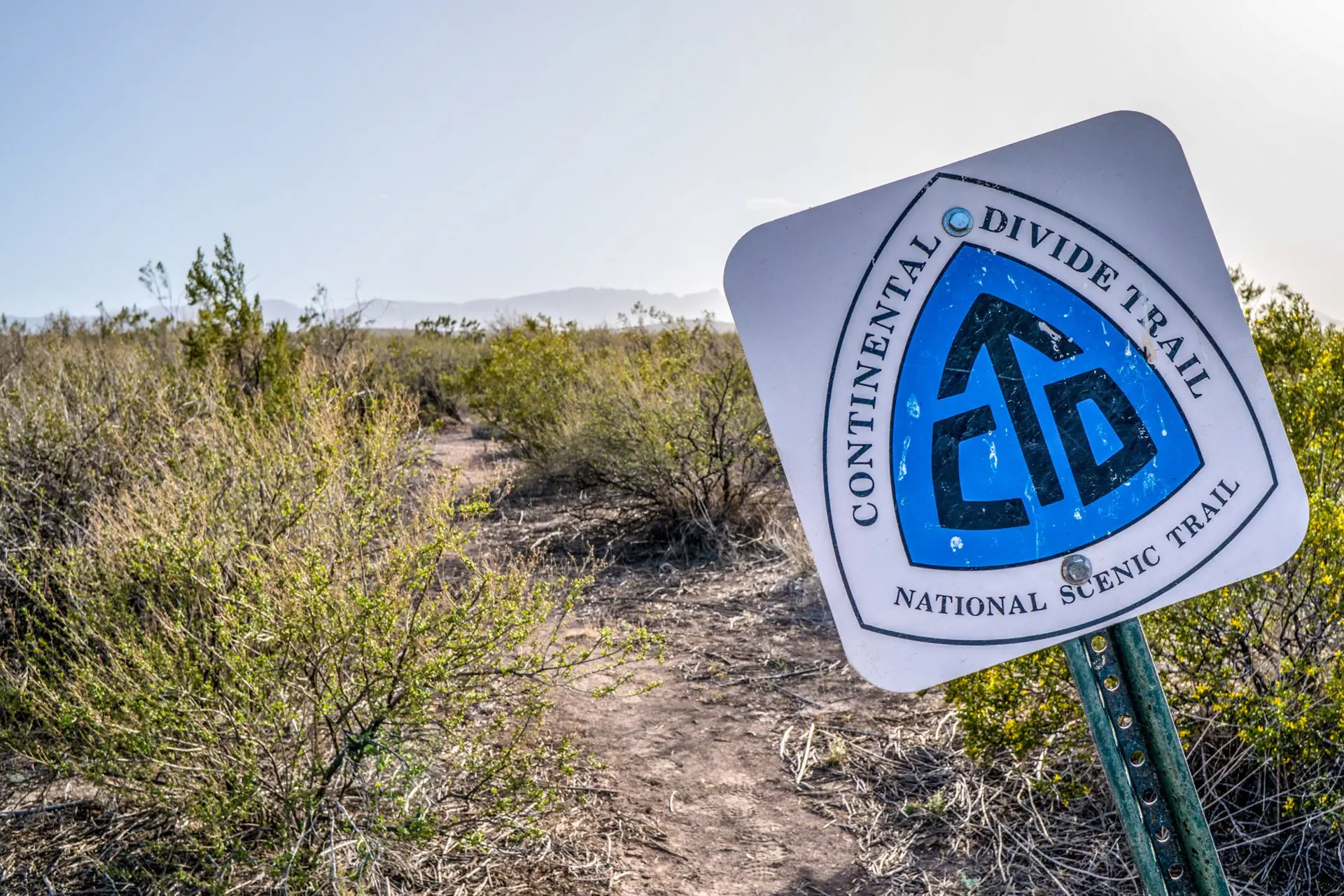 A weathered sign announces the renowned Continental Divide Trail, featuring its iconic logo along a dusty path. Dry bushes and plants border the trail beneath a clear blue sky, with distant mountains providing a breathtaking backdrop.