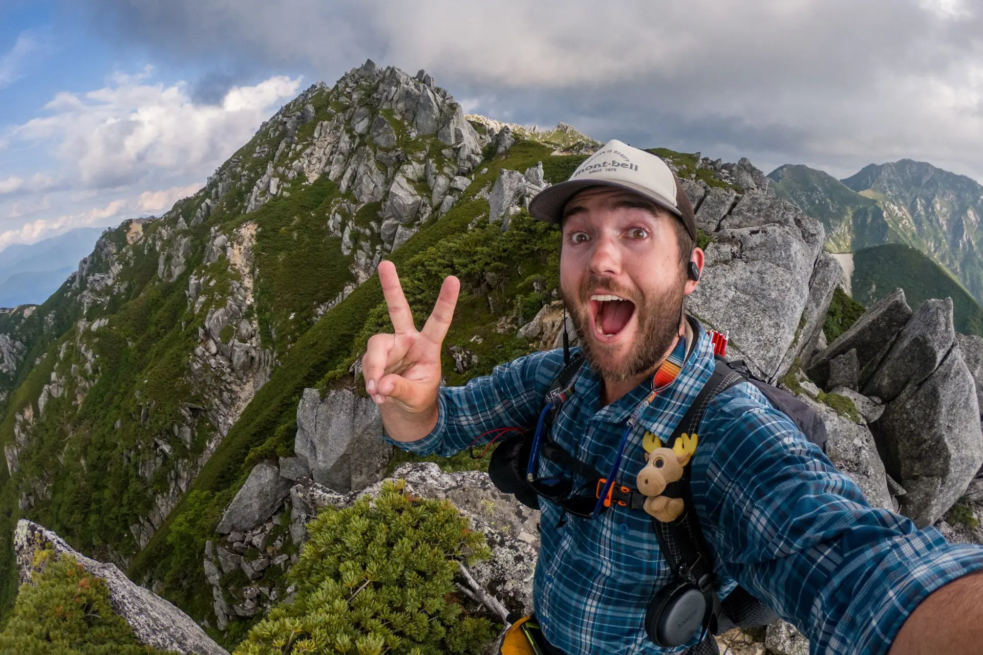 A cheerful man takes a selfie on a rugged peak during his Japan Alps traverse, flashing a peace sign. Clad in a blue plaid shirt and cap, he sports a small toy moose on his backpack. The rocky backdrop highlights the mountains under a partly cloudy sky.