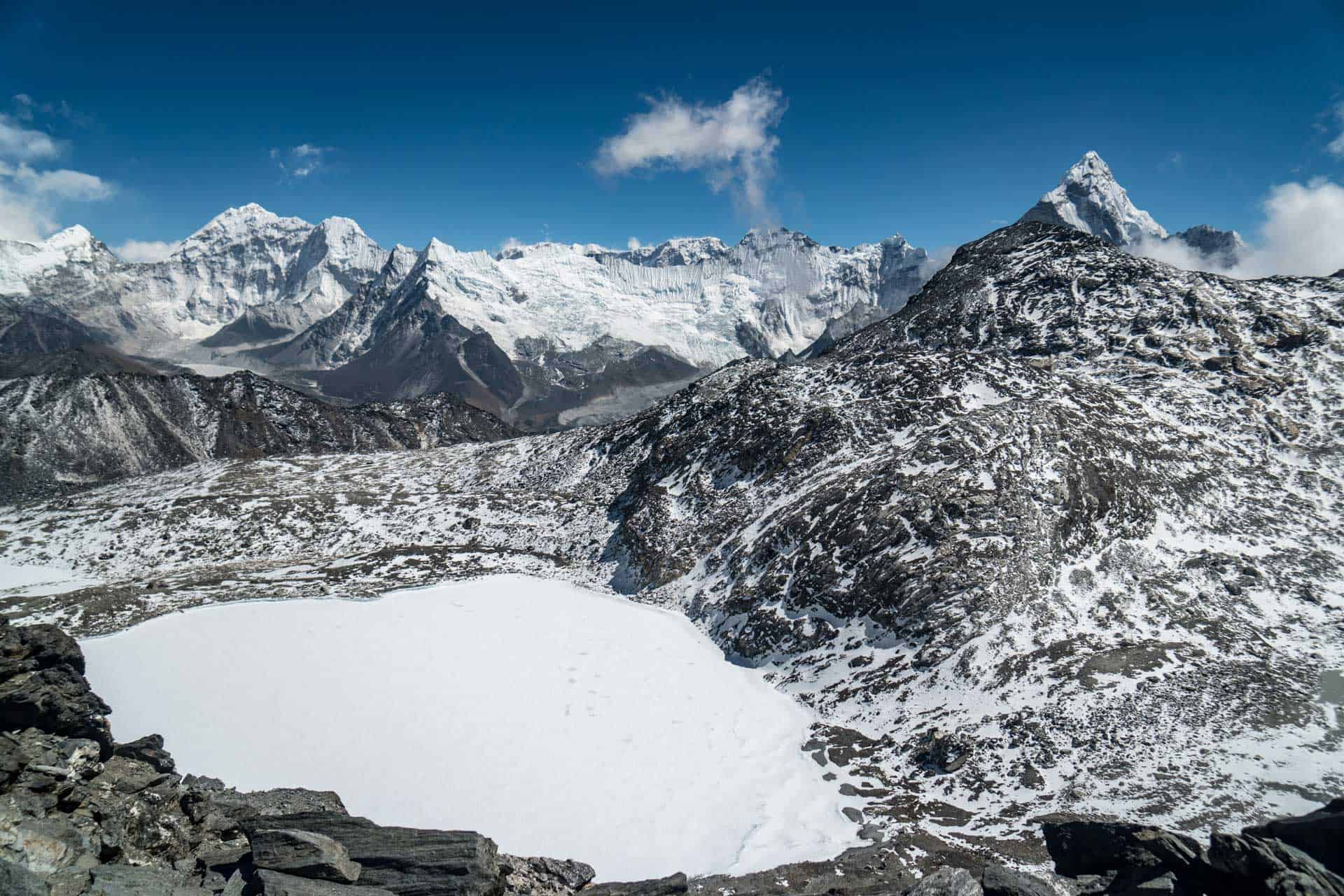 Snow-covered mountains under a clear blue sky surround a frozen lake, reminiscent of the breathtaking views on the three passes trek. Rock formations and patches of snow in the foreground accentuate the rugged terrain, creating a striking alpine landscape.