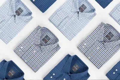 A collection of neatly folded mens shirts, including the Meridian Dress Shirt 2.0, is arranged in a grid pattern. These shirts feature various blue and white checkered designs against a white backdrop, showcasing diverse patterns and styles.