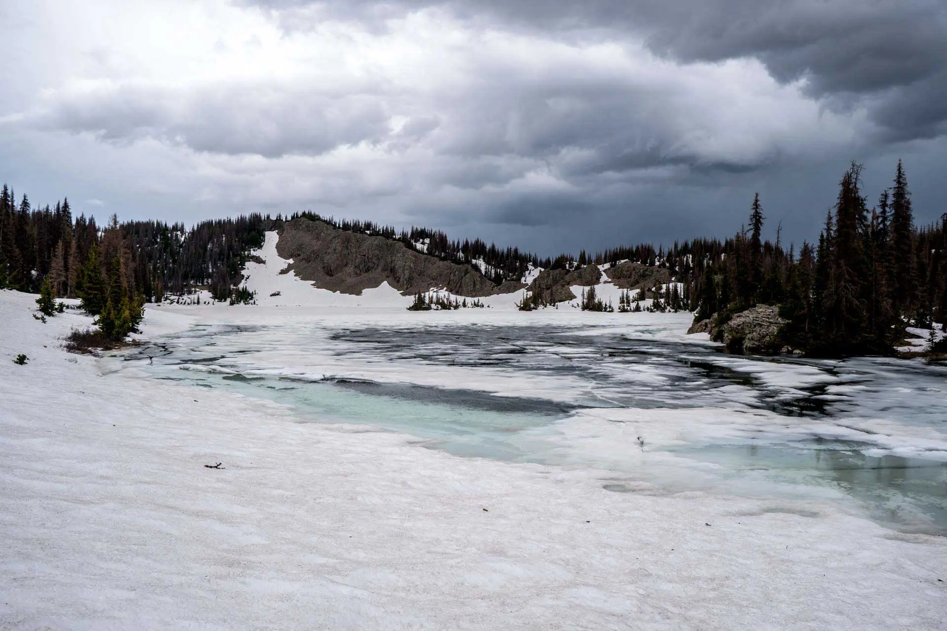 A partially frozen lake nestled in the San Juan Mountains, surrounded by snow and evergreen trees under a cloudy, overcast sky. The scene is serene with a mix of snow and visible water, framed by distant hills.