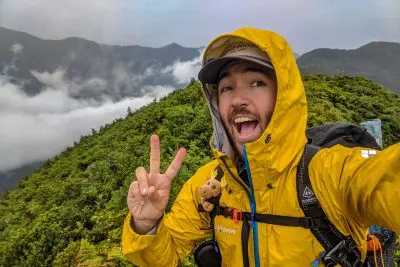 A person in a yellow raincoat and cap, possibly a Montbell Storm Cruiser, is smiling widely and making a peace sign while standing on a lush mountain with fog-covered hills in the background. They carry a backpack with a small plush toy attached.