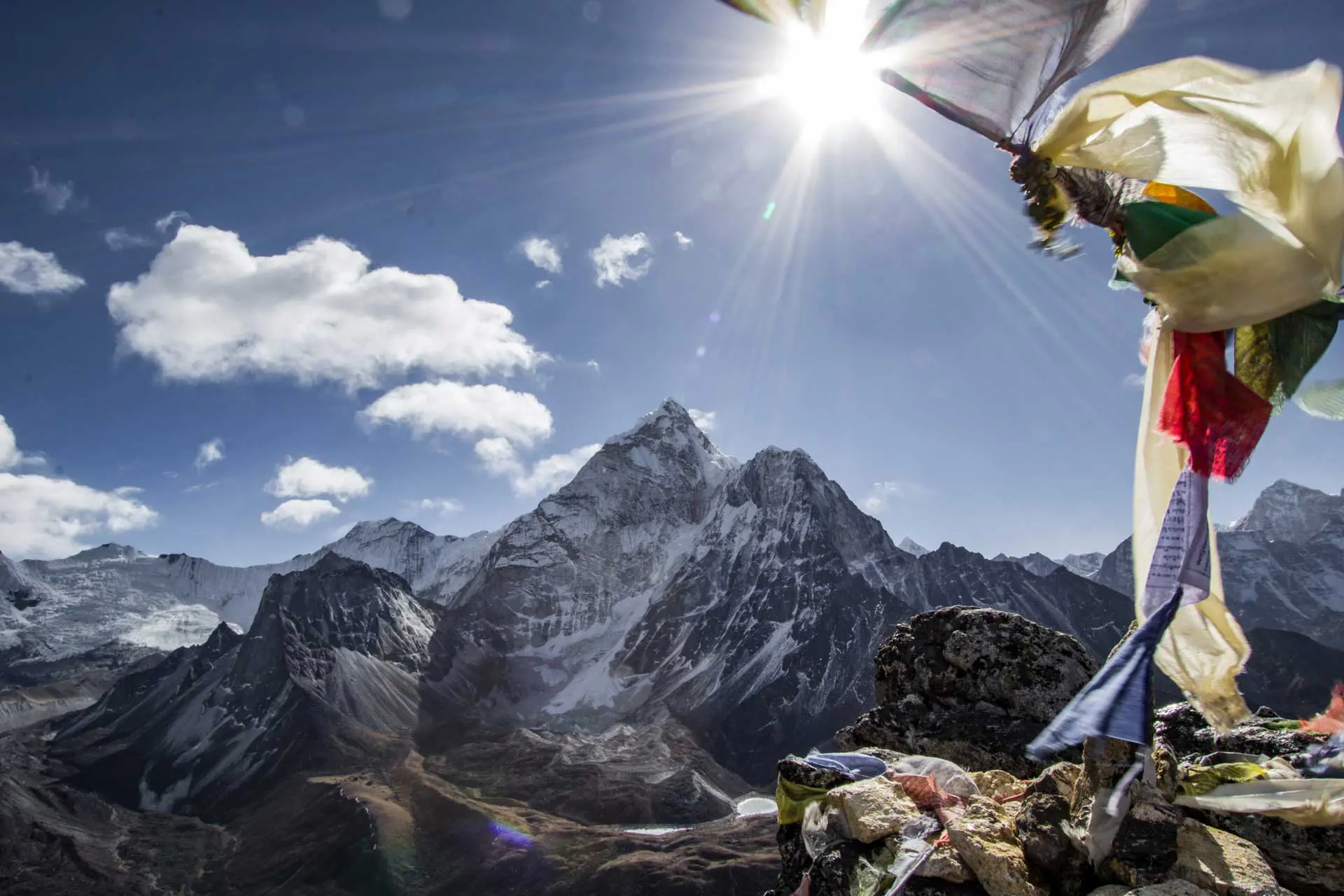 Snow-capped mountains under a bright sun, with clouds scattered in a blue sky. In the foreground, colorful prayer flags flutter on a rocky outcrop, marking the route on the three passes trek map.