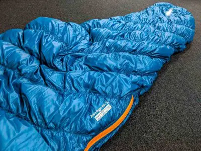 A blue sleeping bag with an orange zipper is spread out on a dark carpeted floor. Labeled Montbell Down Hugger 900 #3, it boasts a cozy quilted design, perfect for your next adventure.