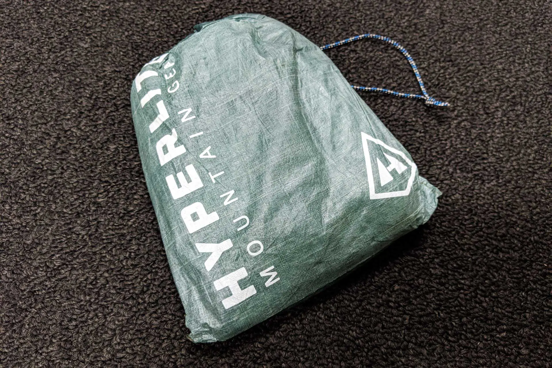 A green drawstring bag from Hyperlite Mountain Gear, perfect for storing your essentials or even a ground cloth, rests on a dark textured surface. The bag proudly displays its logo and brand name in crisp white lettering.