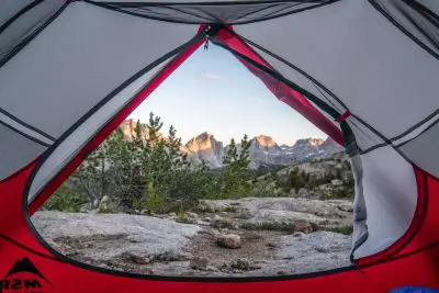 The view from inside the MSR Mutha Hubba NX tent is breathtaking, with the partially open flap framing a picturesque mountain range. Rocky terrain and lush greenery lead to rugged peaks under a clear blue sky, promising an unforgettable outdoor adventure.