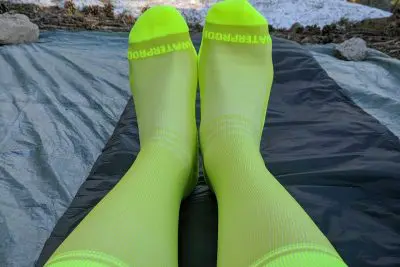 Two feet clad in bright neon green Showers Pass Crosspoint Waterproof Socks rest on a dark-colored tarp outdoors, with rocks and snow framing the background.