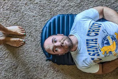 A man relaxes on a blue inflatable pillow, possibly a Therm-a-Rest NeoAir UberLite, on a carpeted floor, gazing upward. Clad in a gray UC Santa Cruz t-shirt with a banana slug motif, he is accompanied by two feet with painted toenails visible nearby.