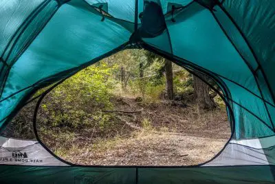 From inside the cozy embrace of a Half Dome 3 Plus tent, the open flap reveals a serene scene: a dirt path winding through lush green foliage and tall trees, perfectly capturing the essence of peaceful camping in the woods.
