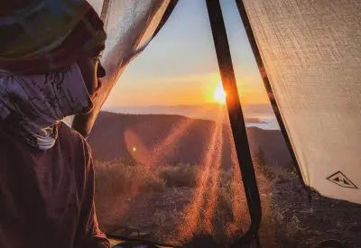 A person wearing a headscarf sits inside a Hyperlite Mountain Gear Dirigo 2 tent, looking out at a vibrant sunrise over distant hills and a lake. The sunlight filters through the tent, casting a warm glow inside.