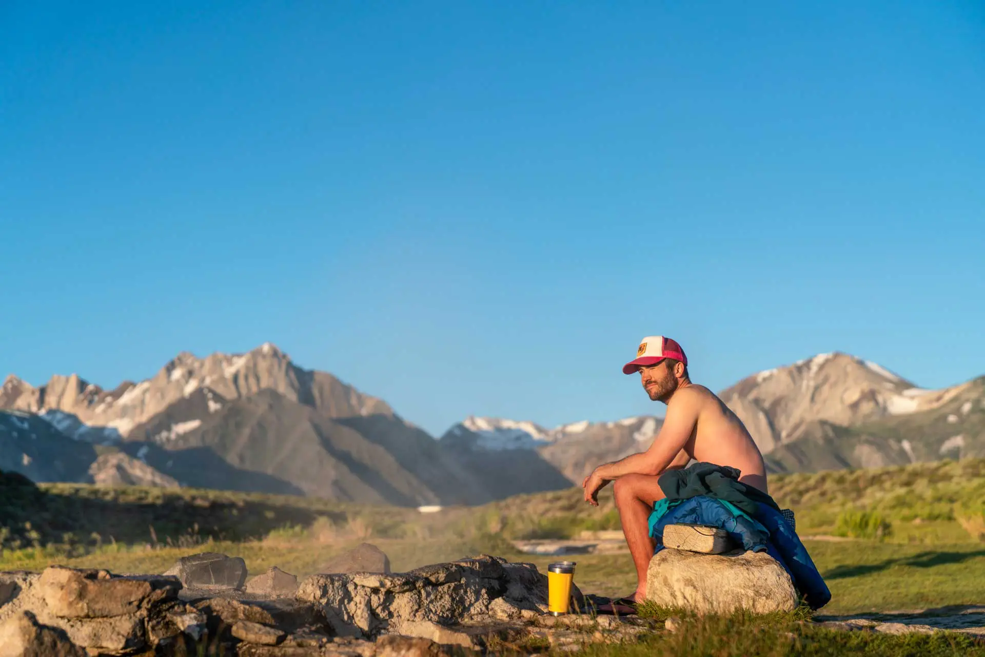A shirtless man, embodying the 2020 agenda of reconnecting with nature, sits on a rock beside a small campfire. He wears a red and white cap and is surrounded by mountains under a clear blue sky, with a blue jacket and yellow mug by his side.
