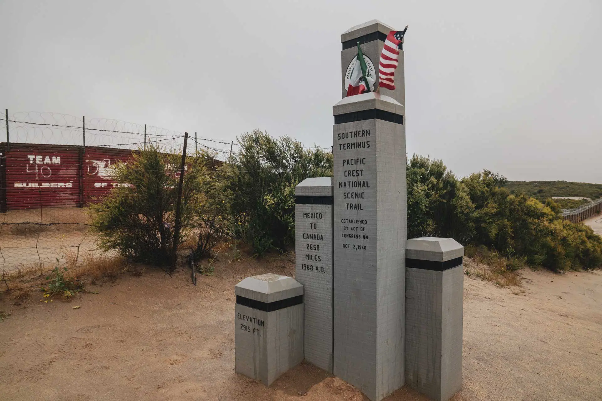 Tall monument marks the southern terminus of the Pacific Crest National Scenic Trail, offering PCT advice on distances to Canada and elevation. An American flag and trail marker crown it, while desert shrubs and a fence with red signs provide a striking background.