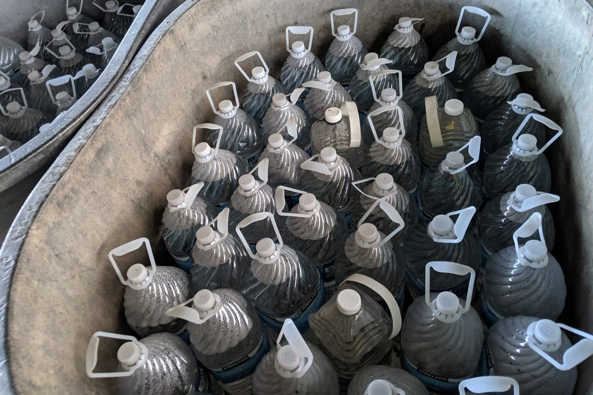 Plastic jugs with white handles, filled with a clear liquid, are neatly arranged like water caches in large, round containers. Lined with a light-colored material, the jugs boast ridged sides and sealed caps.