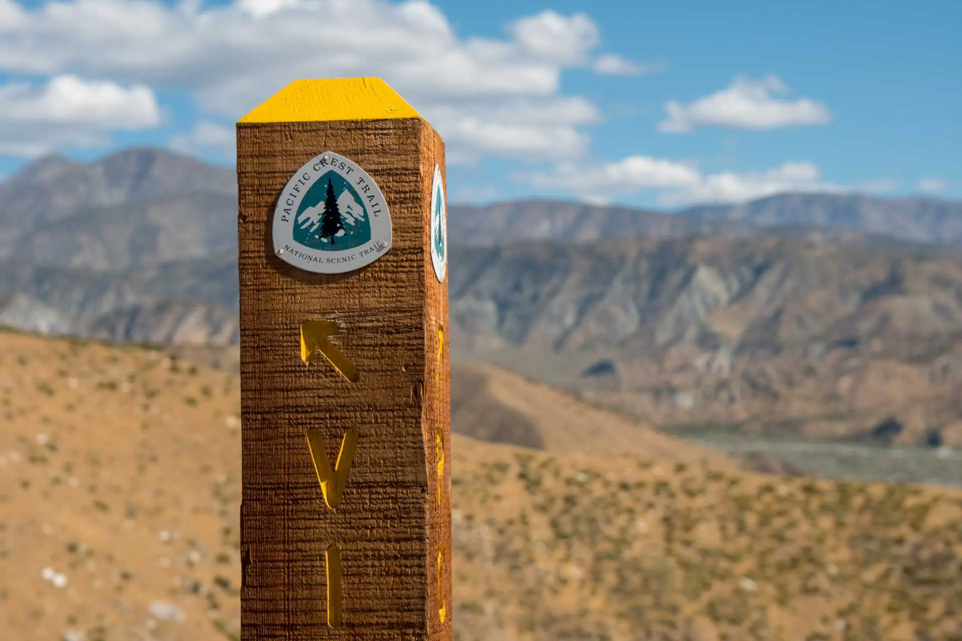 A wooden post with a Pacific Crest Trail sign stands against a backdrop of rugged mountains and a cloudy sky, marking the path for those embarking on the Pacific Crest Trail survey in this dry, rocky landscape.