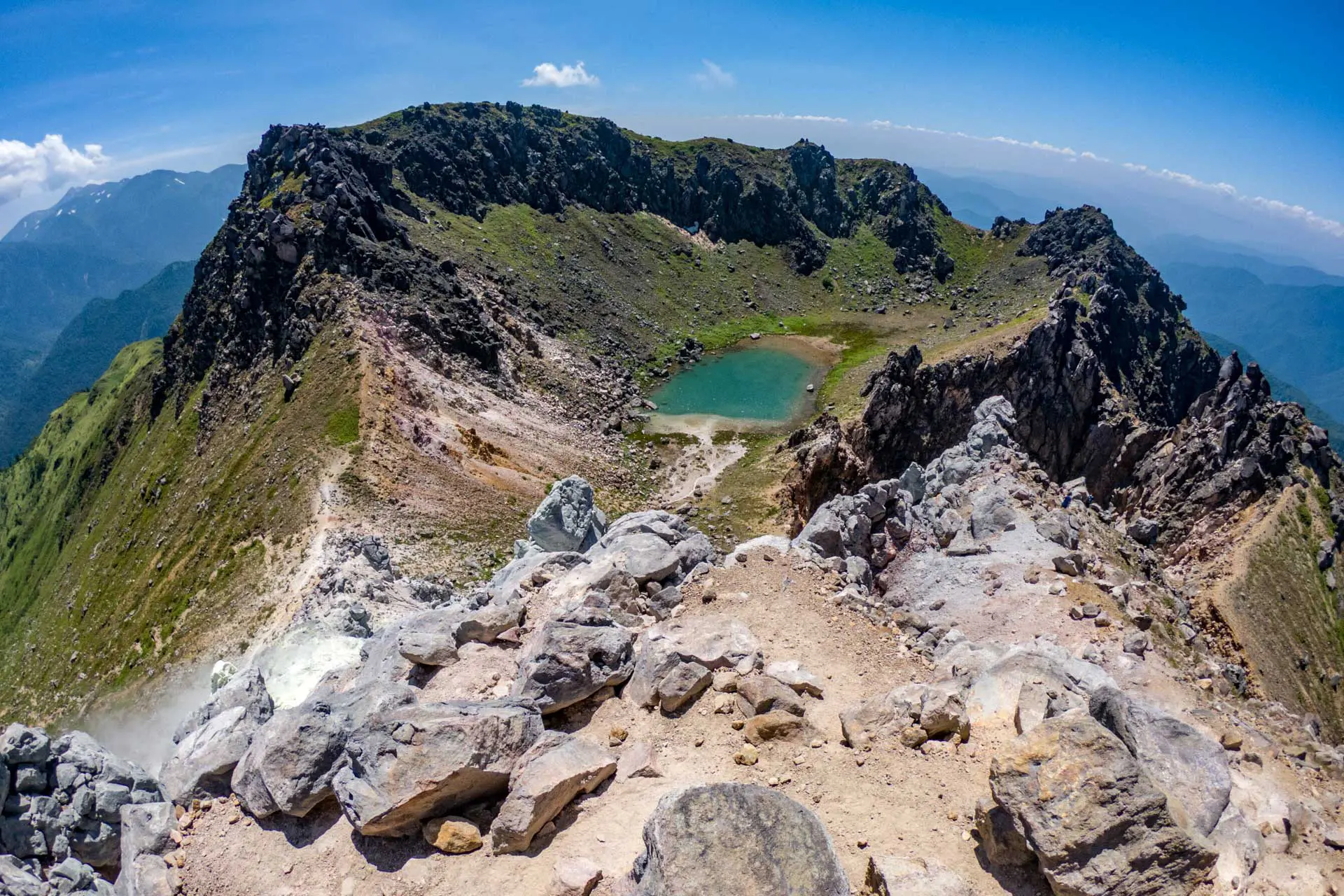 The rocky mountain summit at Yakedake features a rugged path leading to a small green lake nestled in the valley. The surrounding peaks are lush and green under a clear blue sky.