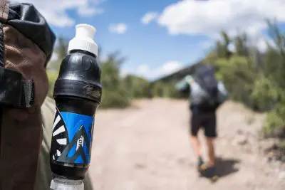 Close-up of a hikers backpack with a water bottle and Sawyer Squeeze filter attached, featuring a blue design. In the background, a person walks on a trail surrounded by shrubs under a partly cloudy sky.