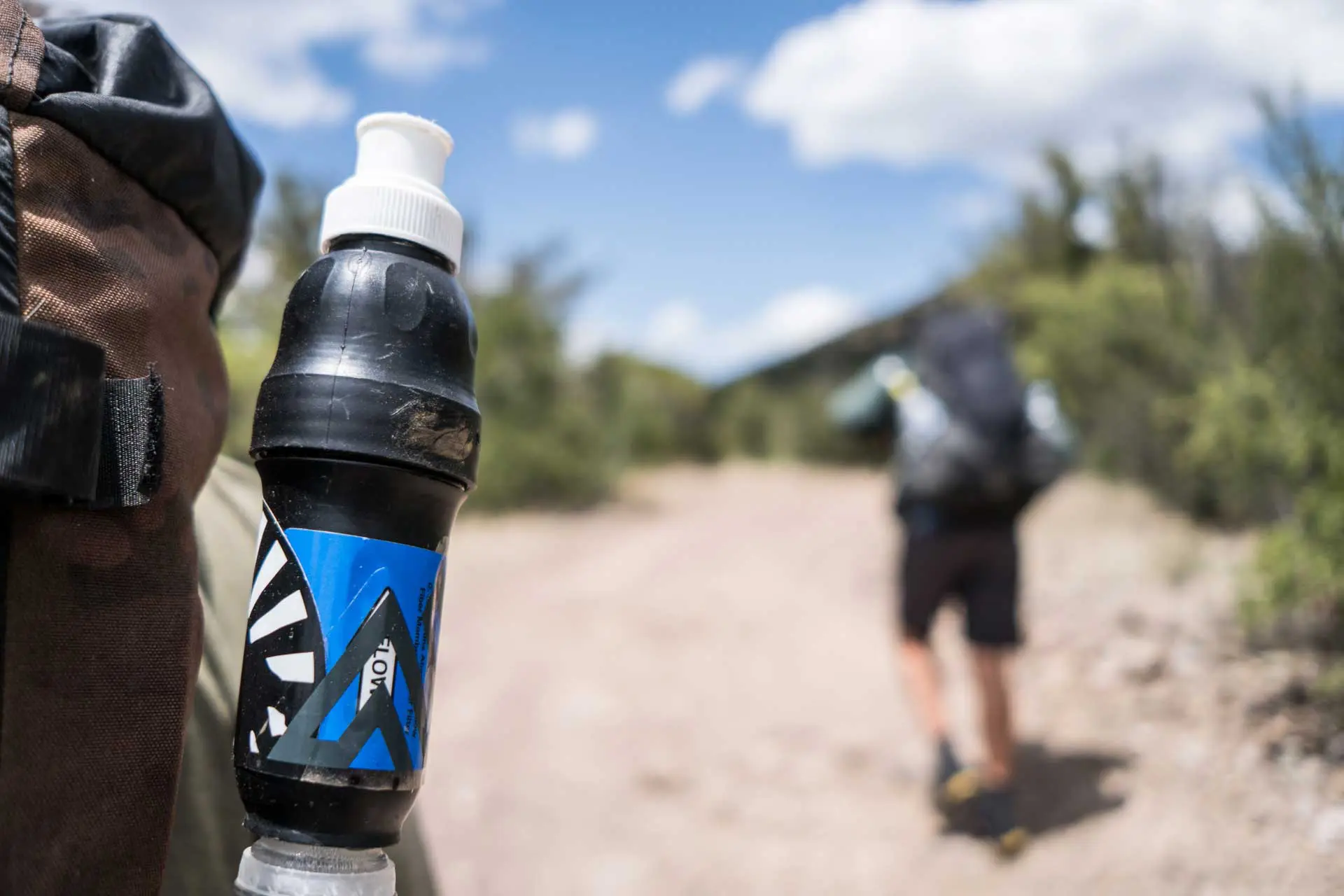 Close-up of a hikers backpack with a water bottle and Sawyer Squeeze filter attached, featuring a blue design. In the background, a person walks on a trail surrounded by shrubs under a partly cloudy sky.
