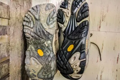 Two Merrell Moab 2 Ventilator hiking shoe soles rest side by side on a wooden and tiled surface. The left sole is worn and dirty, while the right remains new and clean with visibly pronounced treads. Both feature a yellow emblem near the center.