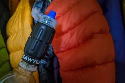 Close-up of a Sawyer Squeeze filter attached to a bottle, set against a backdrop of colorful, puffy jackets hanging side by side.
