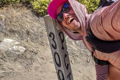 A person wearing sunglasses, a pink hat, and a River Run hoodie smiles widely while leaning next to a wooden post that reads 3000 on a dirt path. Bushes and rocks are visible in the background.