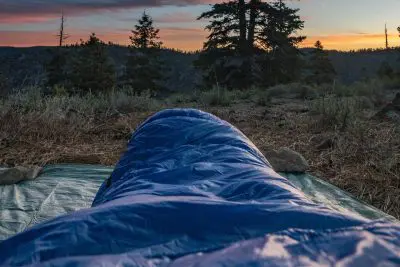 A person lies in a Western Mountaineering UltraLite sleeping bag on the ground, facing a sunset over a forested mountain landscape. The sky glows with hues of orange and pink, while the silhouette of trees stands elegantly in the distance.