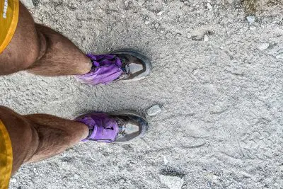 A person stands on a gravel trail, equipped in brown hiking boots with Kahtoola INSTAgaiter gaiters and yellow shorts. The ground is dusty with small stones scattered around.