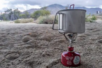 A portable camping stove sits on sandy ground, featuring the MSR PocketRocket 2 and a metal pot. Its fueled by a red canister labeled MSR Isopro. In the background, blurred trees and a mountain rise under a cloudy sky.