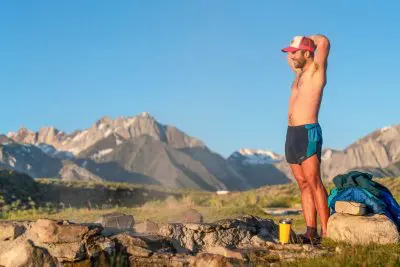A man in Patagonia Strider Pro shorts and a cap stands near a stone campfire ring, stretching with arms overhead. He is surrounded by grass and a mountainous landscape under a clear blue sky. A cup and jacket lie on the ground nearby.