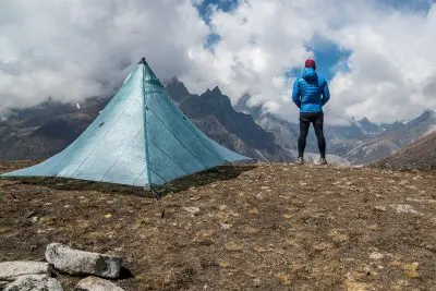 A person in a blue jacket and red hat stands on a rocky plateau next to their Hyperlite Mountain Gear UltaMid 2 tent, overlooking a misty mountainous landscape under a cloudy sky.