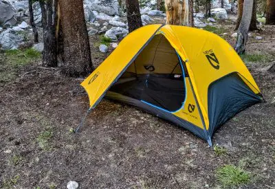 A bright yellow camping tent, the Nemo Hornet Elite 2, is pitched among tall trees on a forested ground, with rocks scattered nearby. The entrance is slightly open, revealing the interior space. The tent proudly displays its logo and name on the sides.