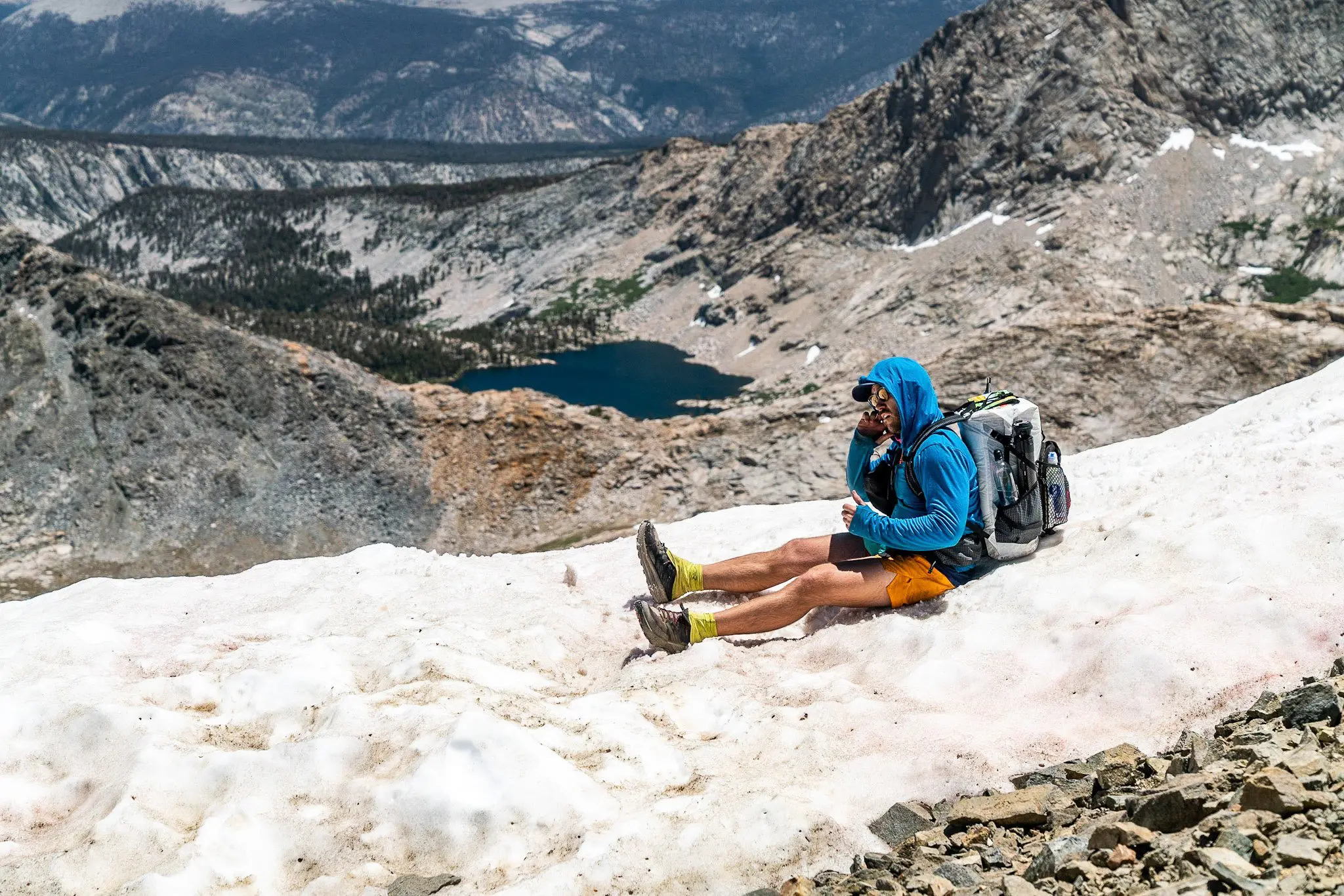 A hiker in a blue jacket and yellow shorts sits on a snowy mountainside, chatting on his phone. With a Hyperlite Mountain Gear Junction backpack and sunglasses, he embraces the adventure. In the background, rocky peaks frame a distant lake beneath a cloudy sky.