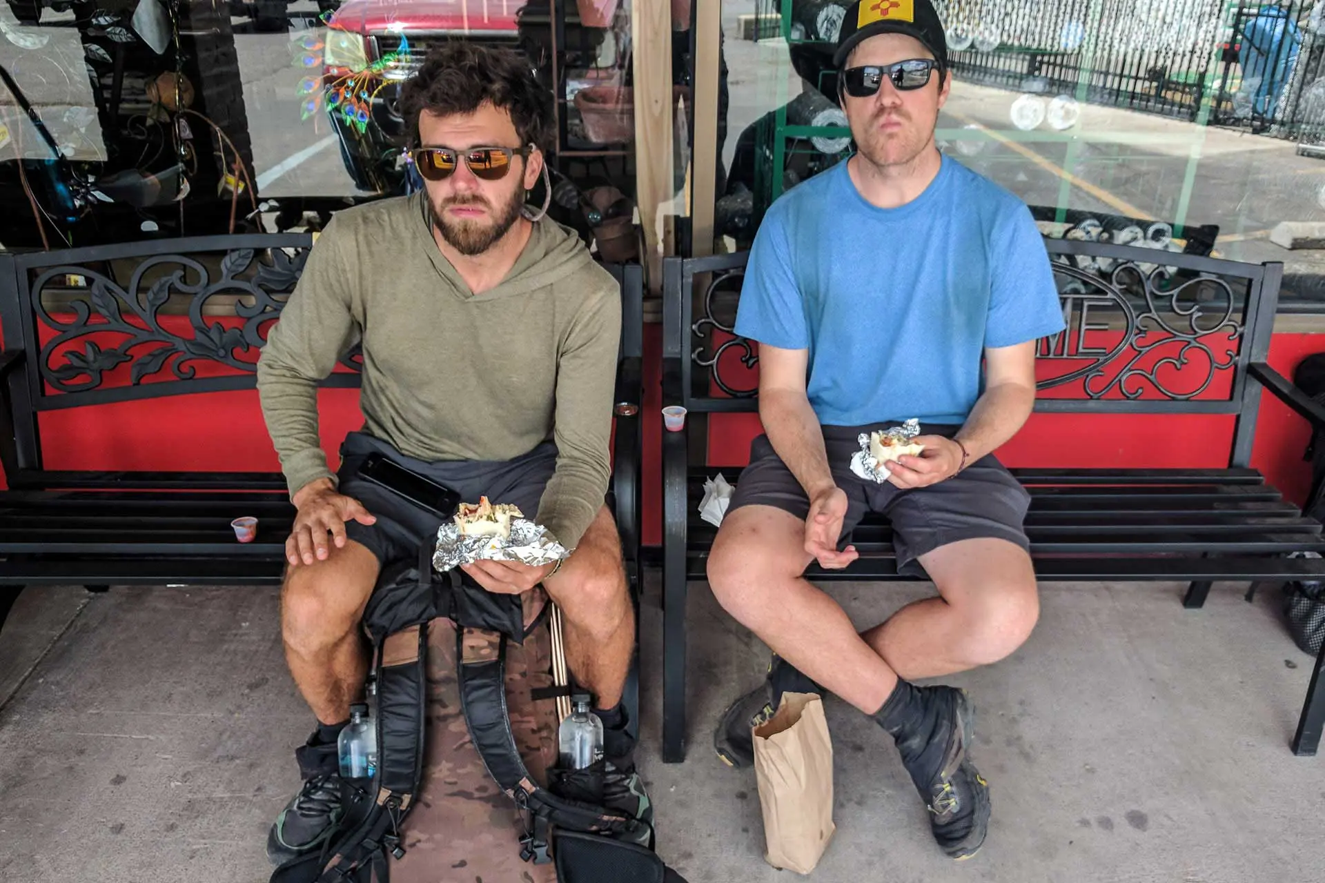 Two men sit on benches outside, eating foil-wrapped food. Both wear sunglasses and casual clothes, and one has prosthetic legs. As they focus intently, it’s almost as though theyre taking in the scene like a cdt survey, reflecting deeply next to a red wall with decorative elements.