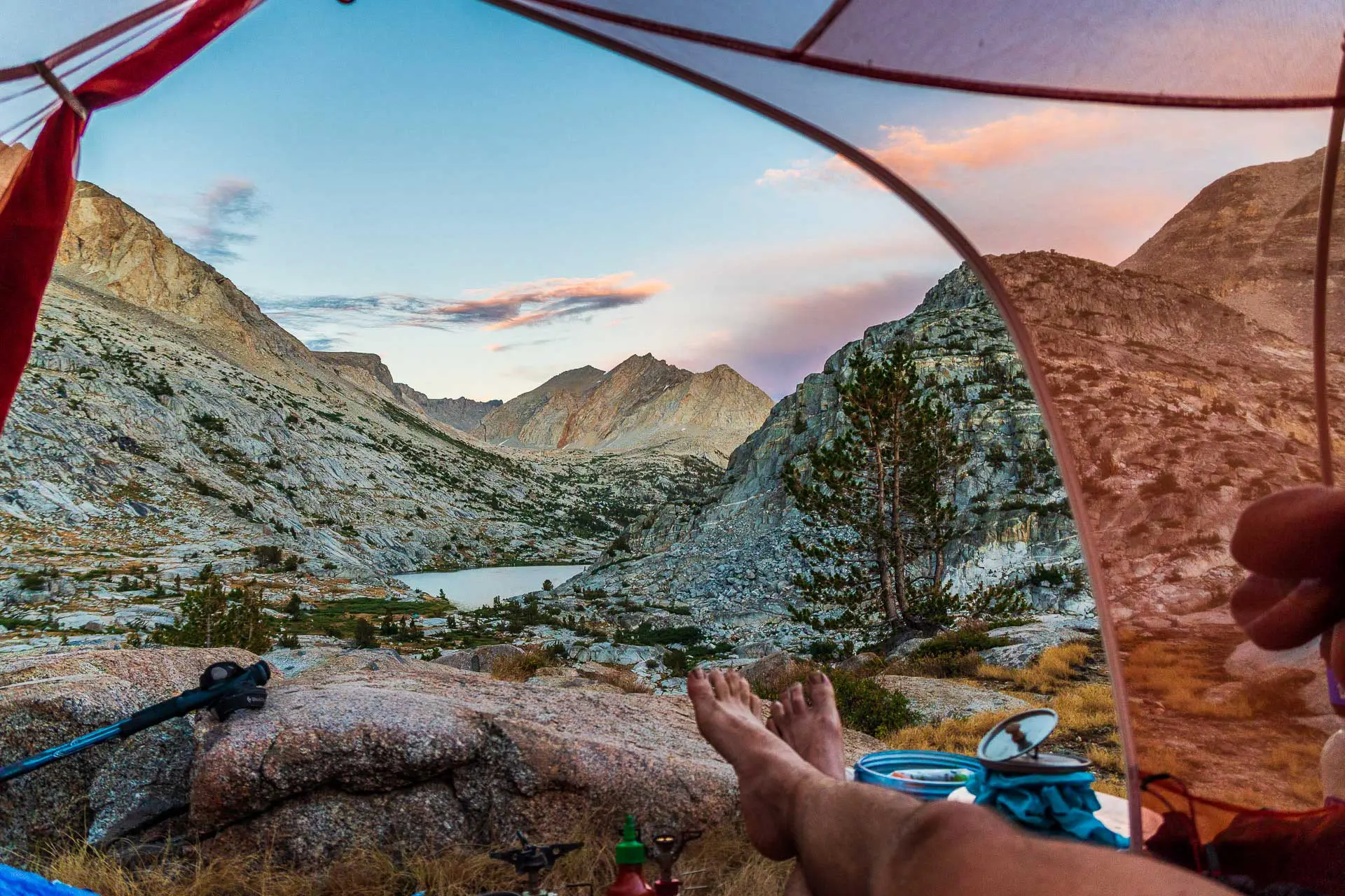 View from inside a tent showcasing a scenic mountain landscape, with a person’s legs stretched out in the foreground. The rocky terrain, small lake, and colorful sunset sky are breathtaking, while camping gear from the PCT Gear Guide 2020 is visible near the tent entrance.
