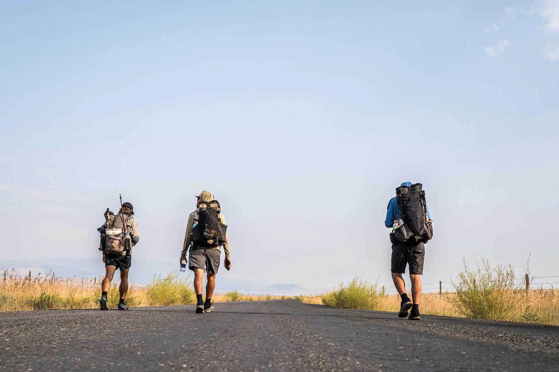 Three hikers with backpacks outfitted with CDT gear walk down an open road under a clear blue sky. The surrounding landscape is flat with dry grass and scattered shrubs. They appear to be trekking or on an adventure, moving towards a distant horizon.
