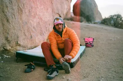 A person sits on a crash pad outdoors in a rocky area, wearing an orange Feathered Friends EOS puffer jacket, red pants, and a cap. They are putting on climbing shoes, with another pair of shoes and a pink backpack nearby.