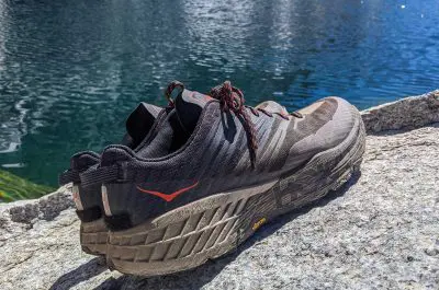 A pair of brown and black Hoka One One Speedgoat 4 trail running shoes with orange accents sits on a rocky surface near a calm body of water. The shoes boast rugged soles, perfect for tackling any outdoor adventure.