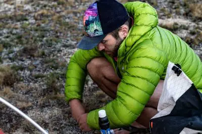 A person in a vibrant green Arcteryx Cerium LT puffer jacket and a colorful cap crouches on the grassy ground, focused on something near their knee. Beside them lies gear including a bottle and a white backpack, all carefully arranged for quick access.