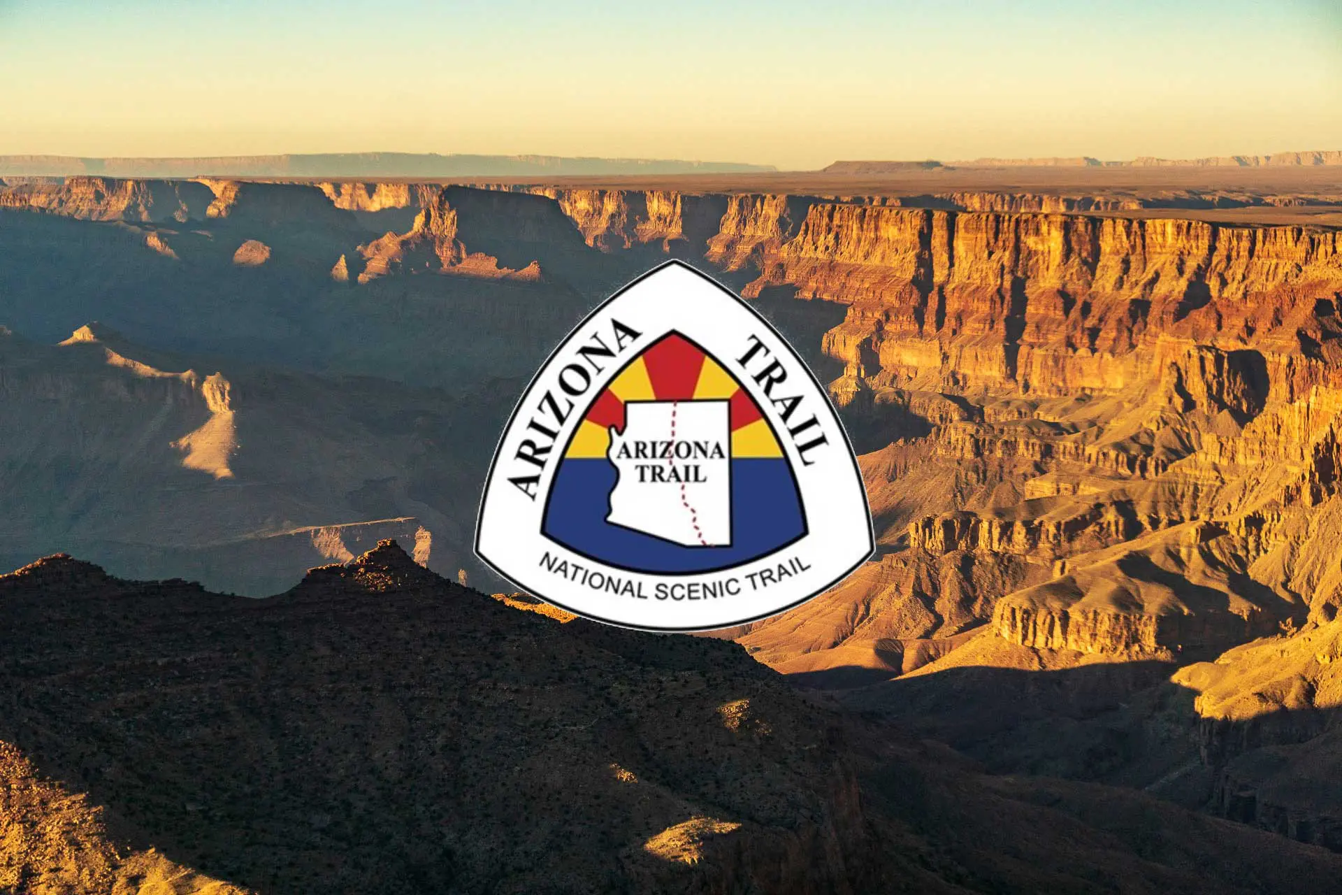 The Arizona National Scenic Trail logo is elegantly superimposed on a breathtaking view of the Grand Canyon at sunset, capturing rocky formations and deep shadows—a perfect backdrop for envisioning a bikepacking adventure along the iconic trail.