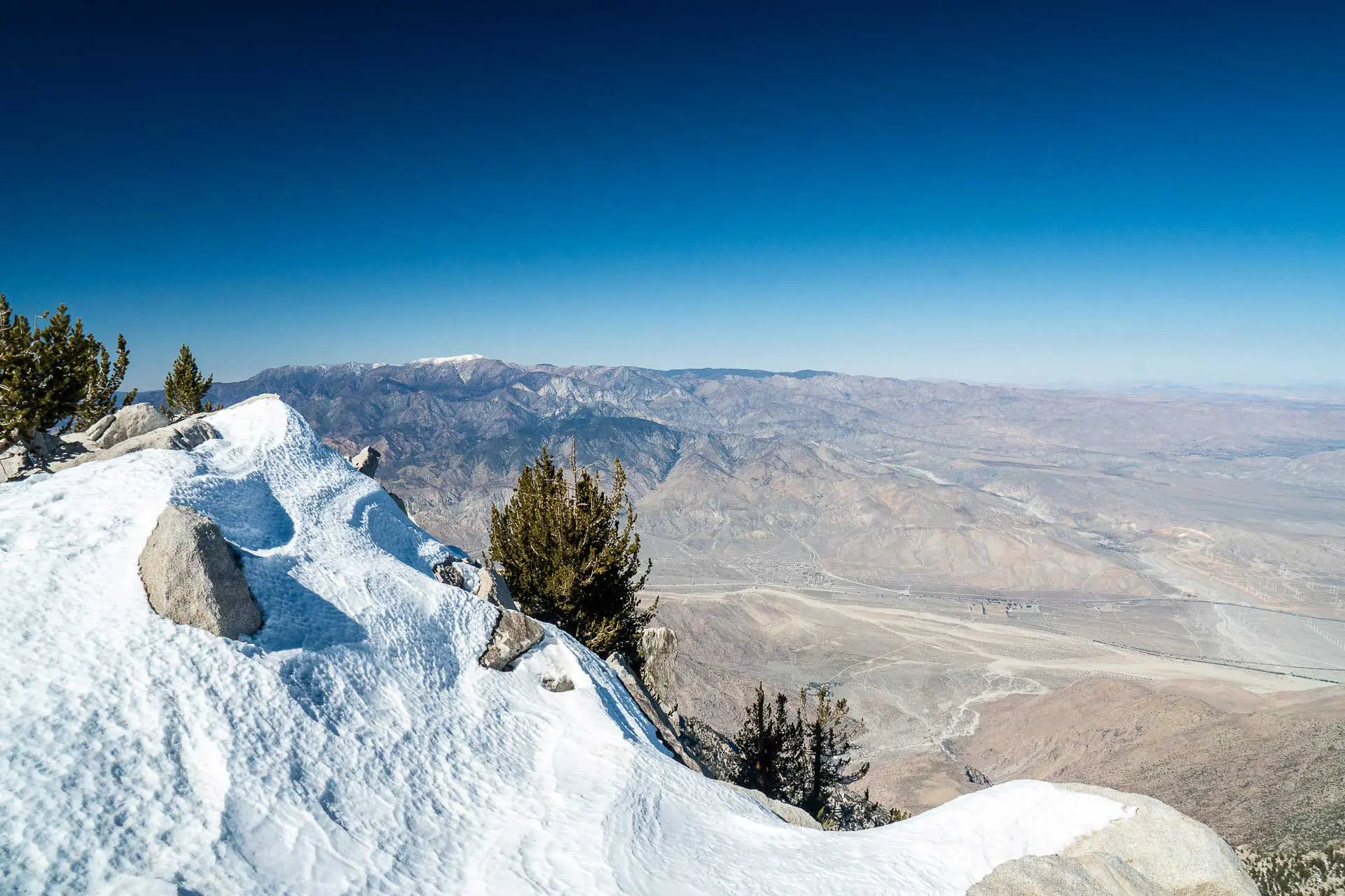 A snowy mountain peak with a few trees overlooks a vast, dry desert landscape under a clear blue sky. The scene evokes the cactus to clouds transition, where rocky terrain contrasts sharply with the white snow in the foreground.