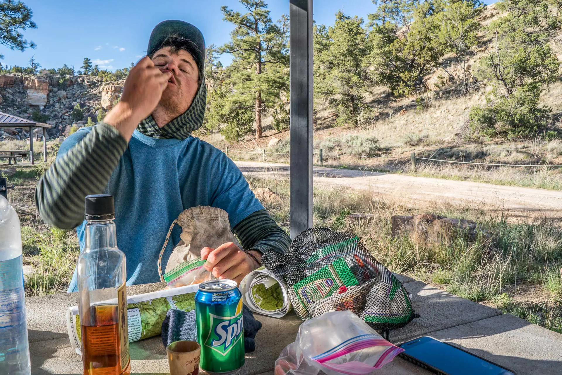 A person in a blue shirt and hat enjoys a CDT resupply moment, eating at a picnic table outdoors, surrounded by trees and hills. Various food items, a can of Sprite, and a bottle cover the table on this sunny day with a clear sky.
